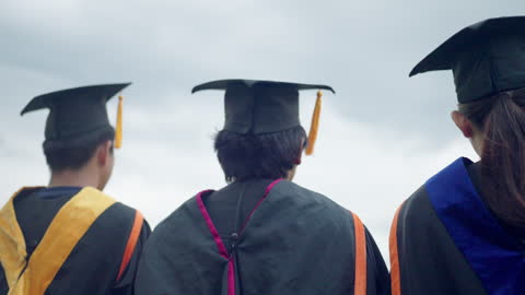 Three graduates are standing in front of a cloudy sky, wearing their caps and gowns. Scene is one of accomplishment and pride, as the graduates have just completed their studies